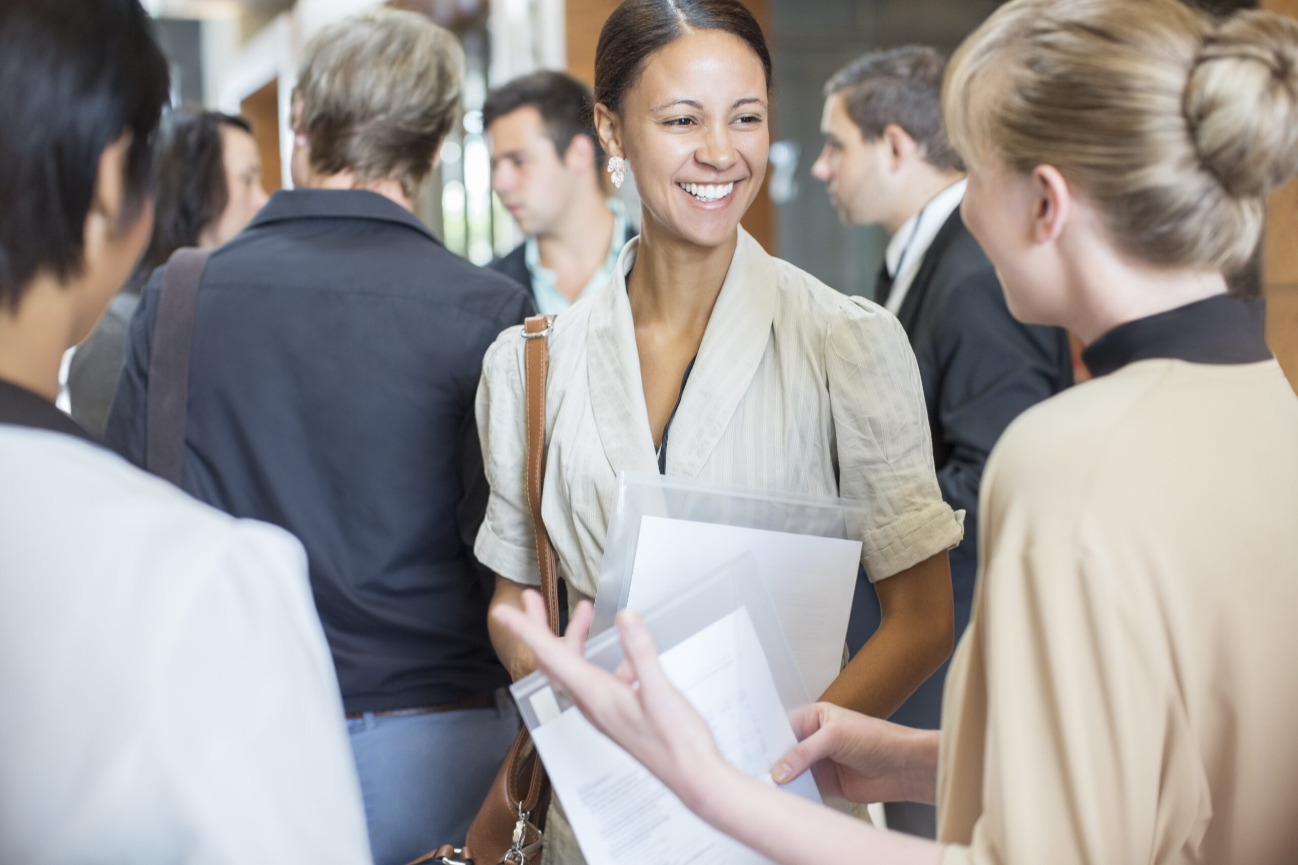 Portrait of two smiling women holding files and talking, standing in crowded lobby