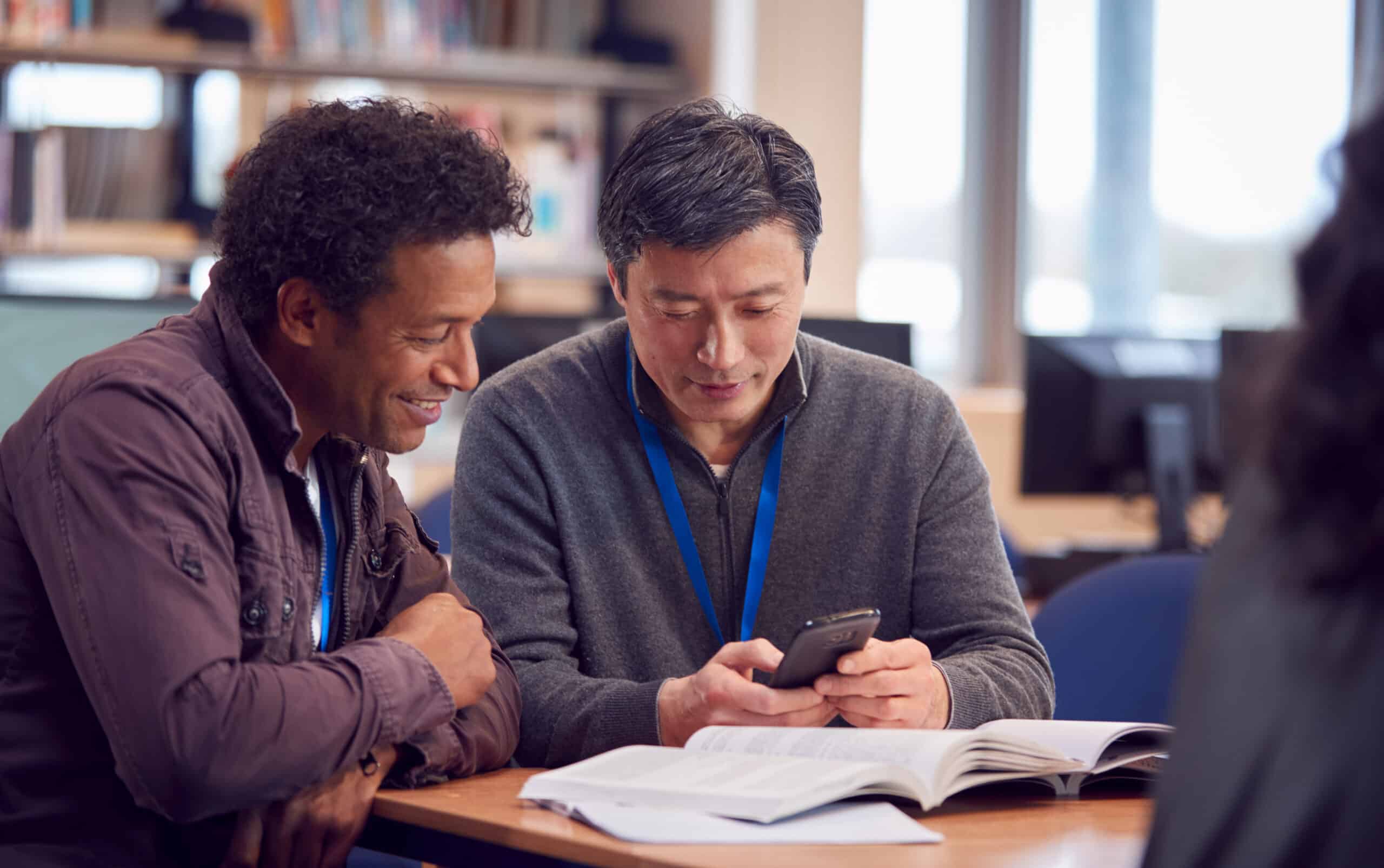 Teacher With Mature Male Adult Student Using Mobile Phone At Table Working In College Library